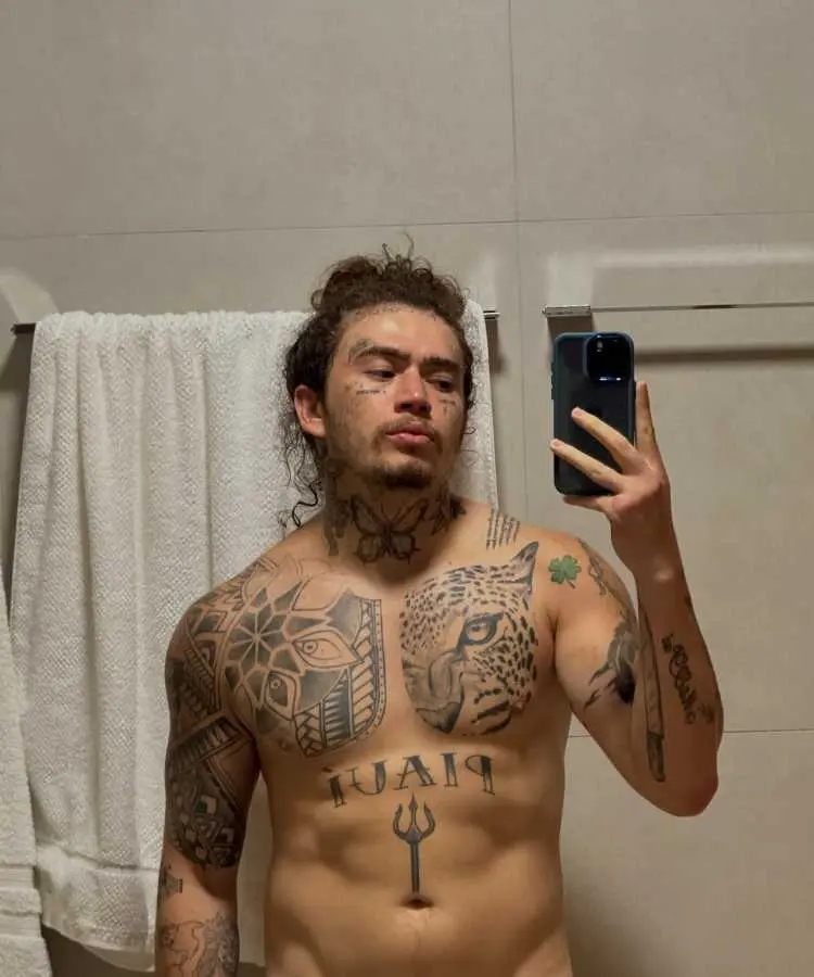 A imagem mostra um homem sem camisa tirando uma selfie em frente a um espelho. Ele possui cabelos presos em coque, várias tatuagens pelo rosto, pescoço e torso, incluindo desenhos detalhados e palavras. Toalhas brancas aparecem ao fundo.