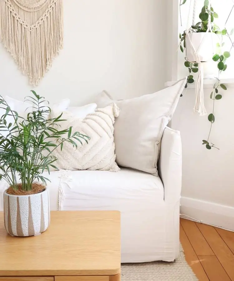 Uma sala clara com sofá branco e almofadas texturizadas. Uma planta verde em vaso listrado está em uma mesa de madeira. Na parede, um macramê decorativo, e à direita, plantas penduradas próximo à janela. O ambiente é acolhedor e com tons neutros.








