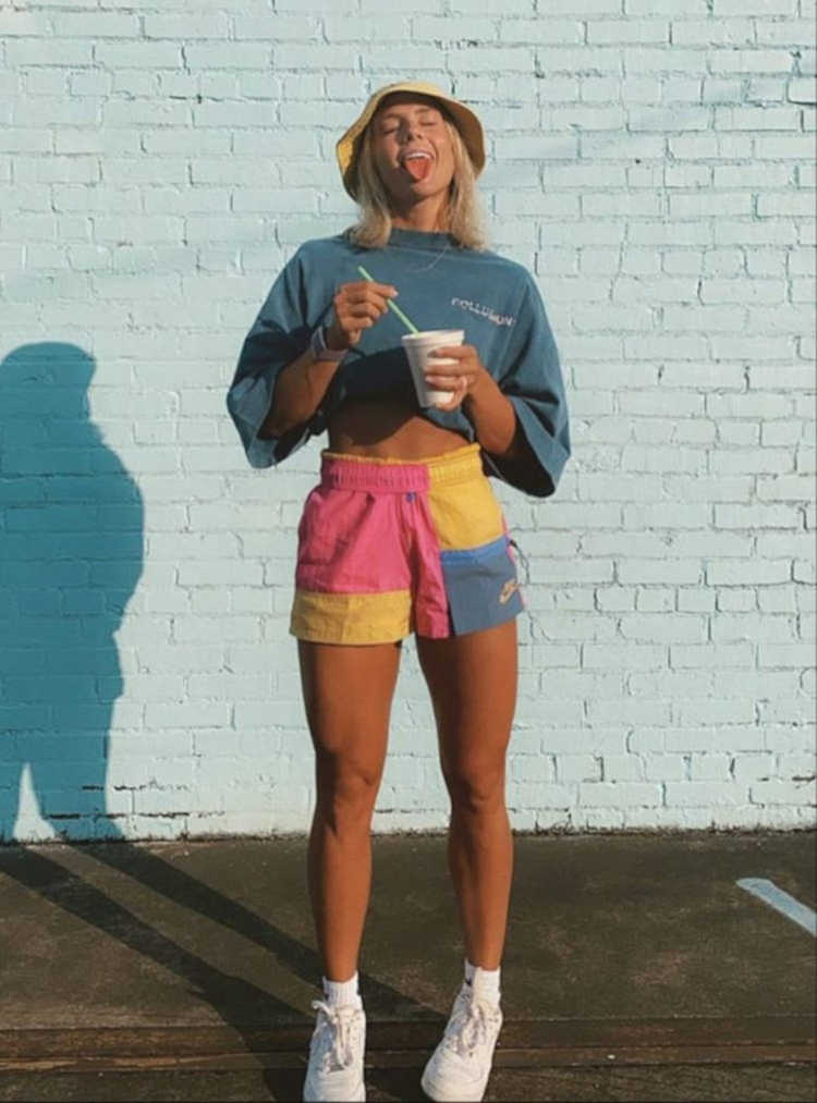 Mulher loira de pele clara usando camisa cropped azil, shorts coloridoe chapéu