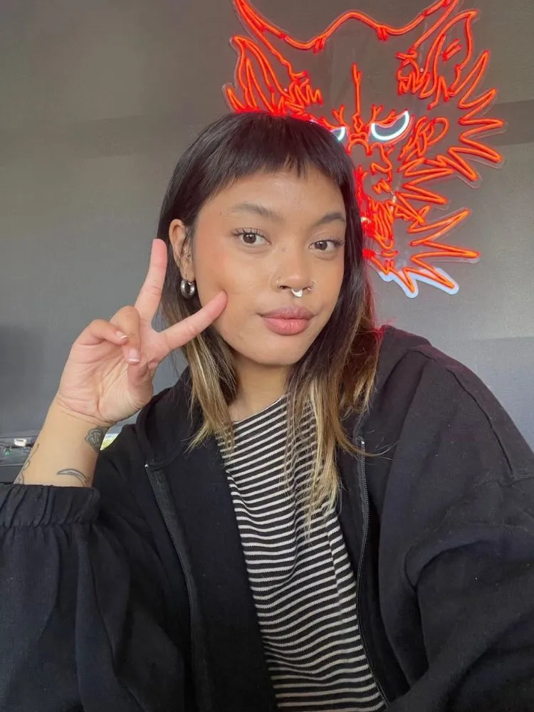 Mulher de pele morena usando blusa branca e preta, casaco preto e corte de cabelo médio com baby bangs