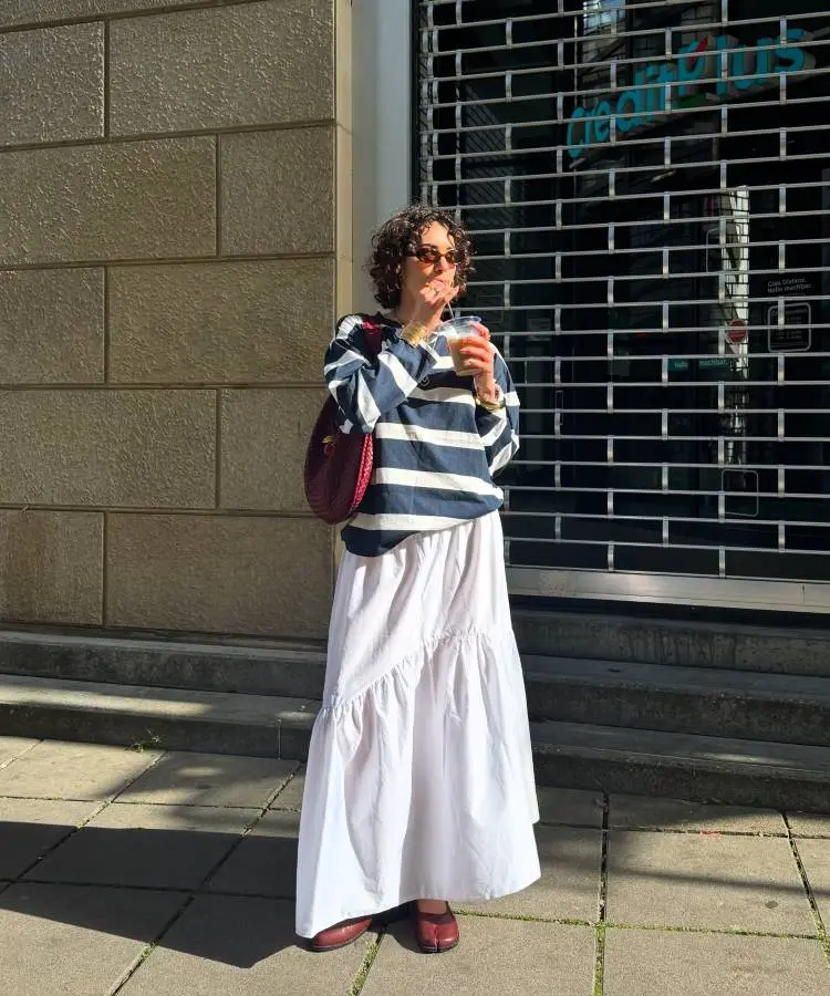 Uma mulher com cabelo cacheado e óculos de sol redondos bebe de um copo enquanto veste uma blusa listrada azul marinho e branca de mangas compridas, combinada com uma longa saia branca esvoaçante. Ela usa sapatos vermelhos e carrega uma bolsa de ombro vermelha.