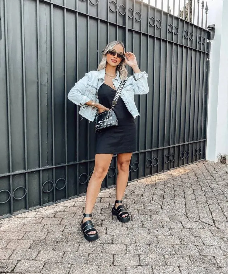 uma mulher loira posa em frente a um portão ela usa um vestido preto, papetes pretas e uma jaqueta jeans