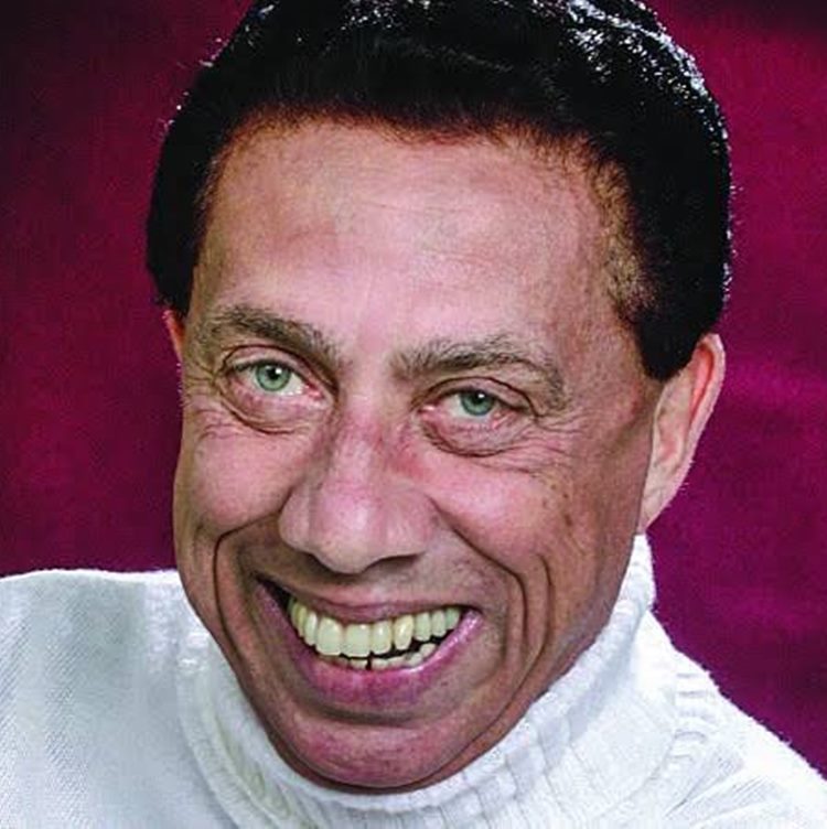 Close-up de um homem de pele morena com cabelos escuros e olhos claros. Ele está sorrindo, mostrando os dentes, e usa uma blusa de gola rolê branca. O fundo é de um tom vermelho.