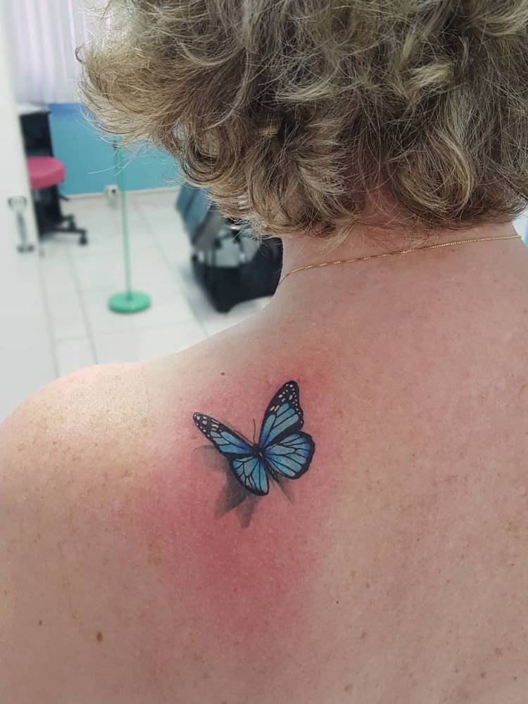 Mulher loira de cabelo curto com tatuagem de borboleta azul sombreada na parte de trás do ombro.