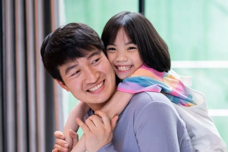 A imagem mostra um pai sorridente, de pele clara e cabelo escuro, carregando nas costas sua filha, também sorridente, com cabelo preto liso. Ambos parecem felizes, olhando na mesma direção, com a menina abraçando o pai por trás.







