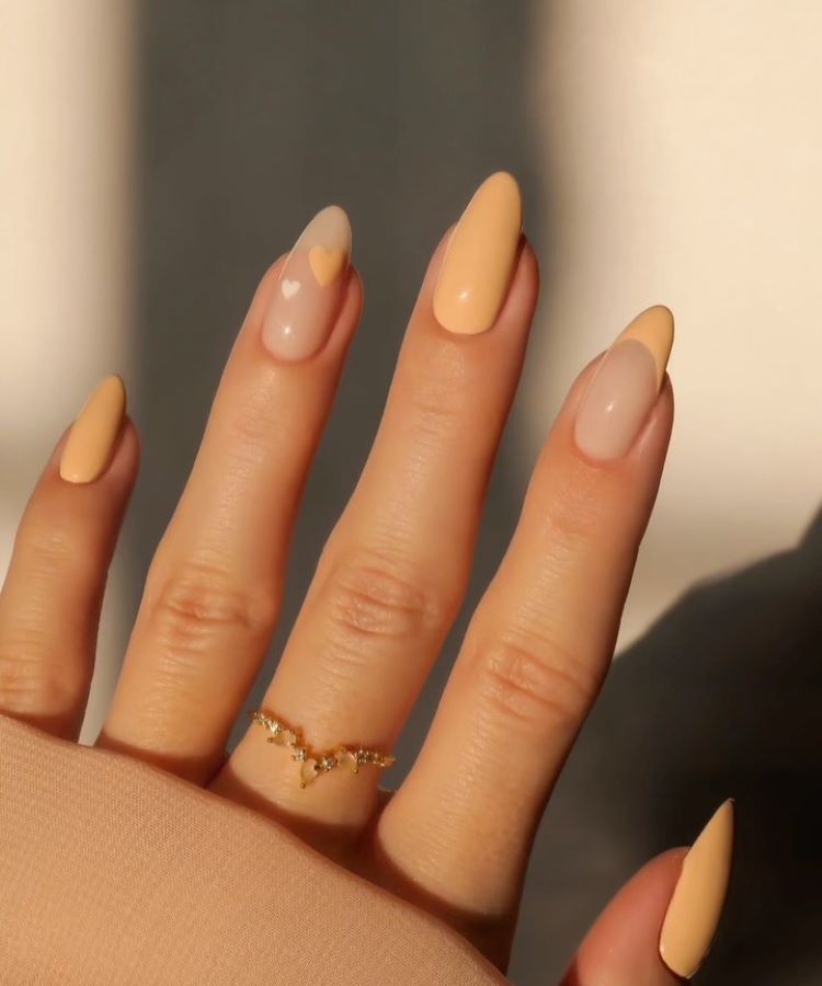 A imagem mostra uma mão com unhas decoradas em tons de pêssego e nude. Duas unhas têm um design de coração, uma delas com um coração pequeno e outra com um coração maior, ambos em uma base translúcida. As outras unhas são pintadas em um tom sólido de pêssego. O anel dourado delicado complementa o visual. Este estilo é um exemplo de unha decorada simples, combinando elegância e sutileza.