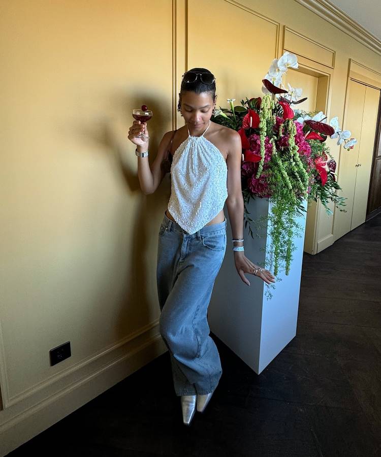 A imagem mostra uma mulher usando uma calça jeans wide leg oversized de lavagem clara, tendência 2025, combinada com um top branco brilhante de amarração no pescoço. O look é finalizado com sapatos metálicos e acessórios discretos, criando um visual sofisticado.