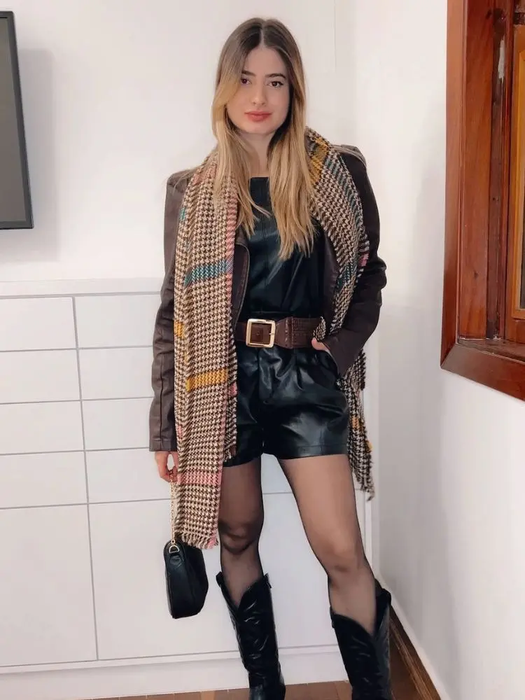 Mulher de pele clara com look de couro preto, jaqueta marrom e echarpe xadrez 