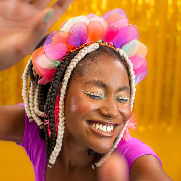 Mulher de pele negra usando roupa roxa, dread no cabelo e tiara com imitação de pétalas coloridas.