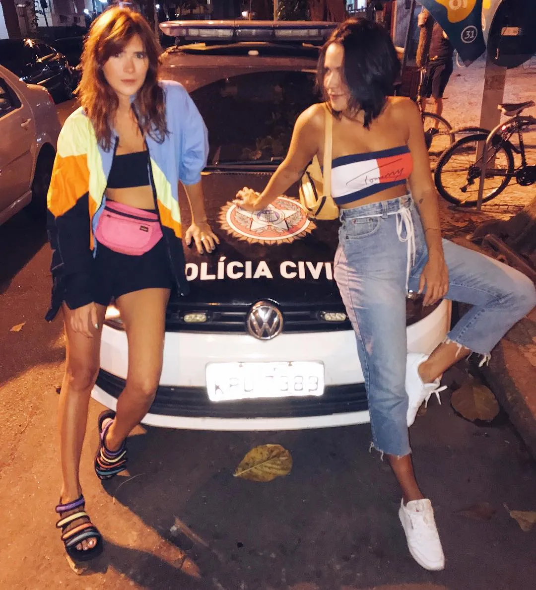 Foto de duas mulheres de pele clara apoiadas em viatura da Polícia Civil. Uma usa top e shorts preto com corta-vento azul, preto, laranja e amarelo e outra usa top azul-marinho, branco e vermelho e calça jeans 