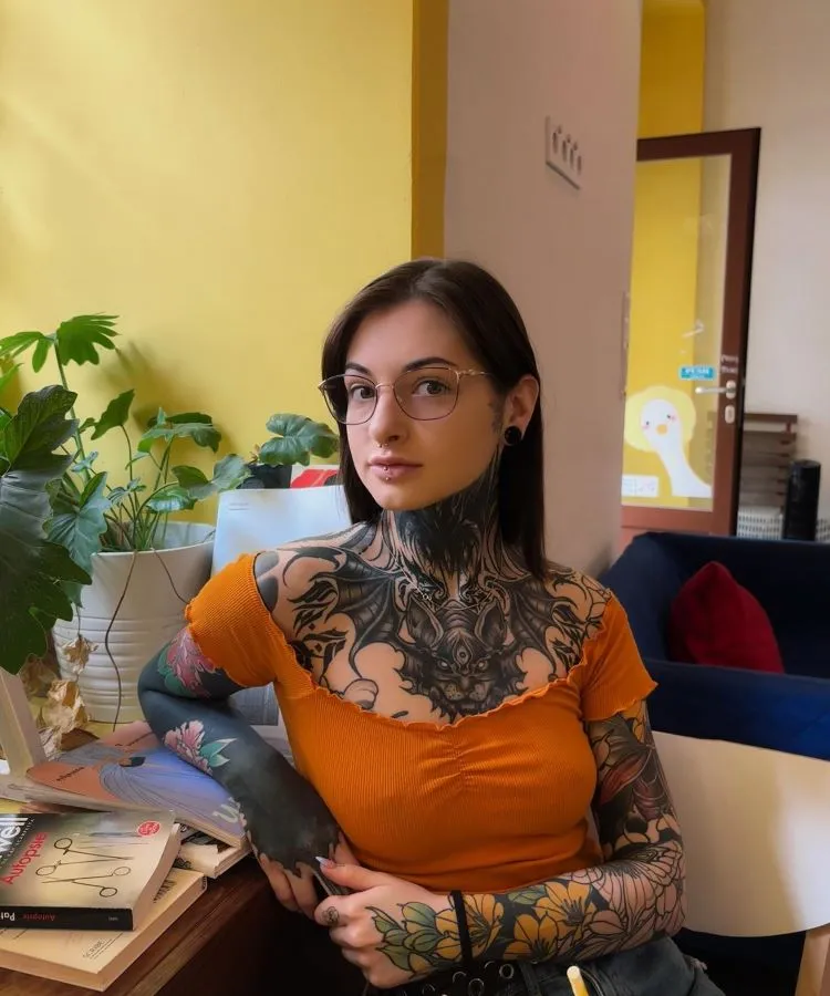 A imagem mostra uma pessoa sentada, com cabelo castanho e óculos. Ela tem uma tatuagem no pescoço que se estende até o peito e ombros, com um design complexo em preto. A pessoa usa uma blusa laranja e está em um ambiente com plantas e livros ao redor.




