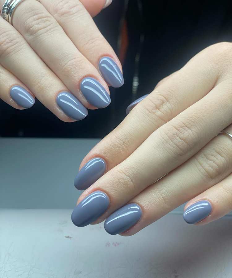 Mãos femininas com unhas no formato amendoado, pintadas em cinza azulado suave e brilhante. O fundo neutro em preto e cinza destaca a delicadeza e a modernidade do esmalte, representando uma das principais tendências de 2025 para unhas elegantes.