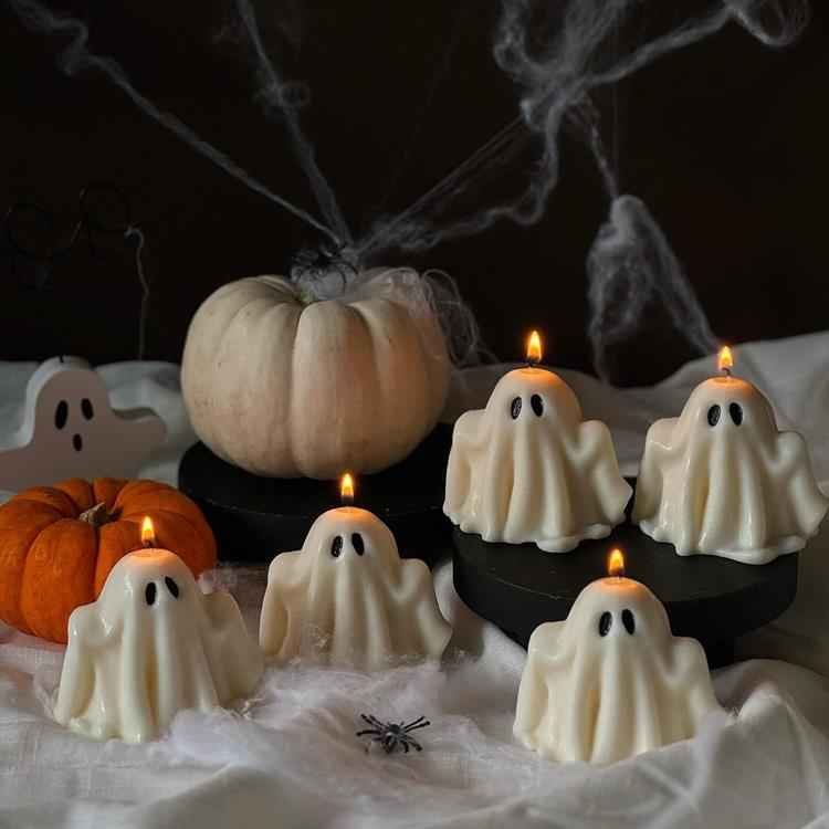 velas acesas e pequenas em formato de fantasma sobre mesa com toalha branca e ao lado de abóbora