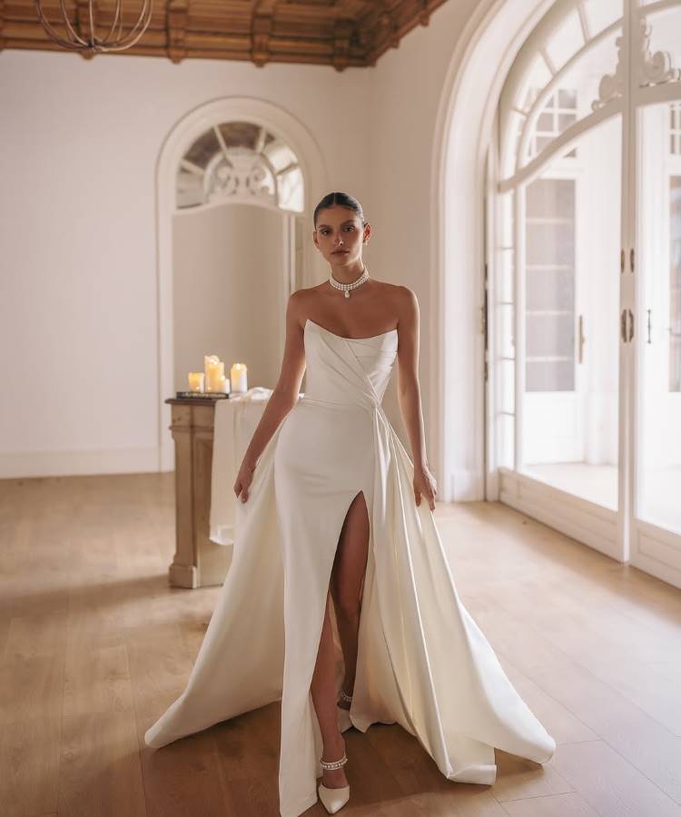 Noiva em vestido branco tomara que caia com drapeado elegante, fenda alta e cauda fluida. Ela usa colar de pérolas, destacando sofisticação. O ambiente possui piso de madeira, teto detalhado e janelas arqueadas com luz suave.





