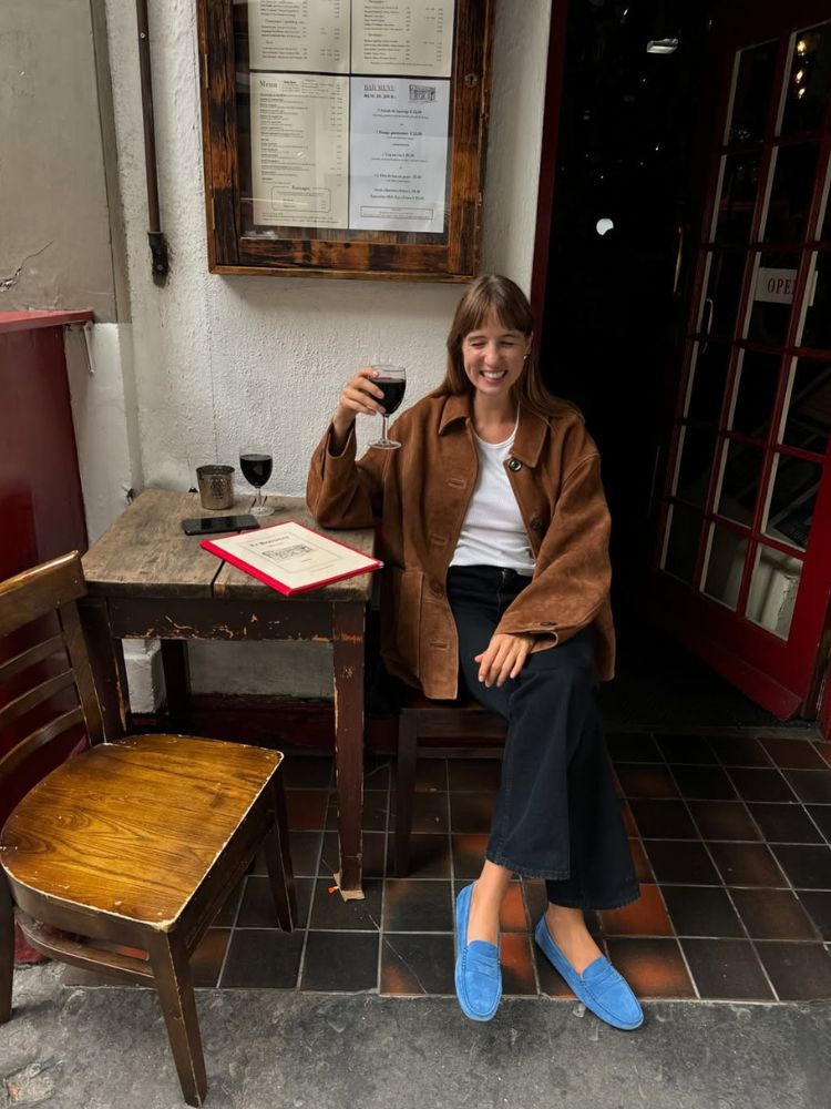 Mulher de pele clara sentada à mesa usando camiseta branca, calça jeans, casaco marrom de camurça e mocassim azul de camurça 