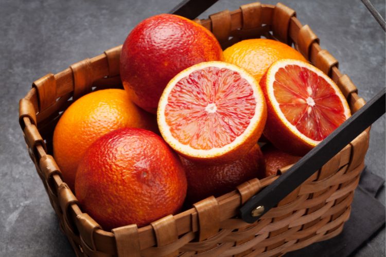 Uma cesta de vime contém várias laranjas-sangue inteiras e cortadas ao meio. As frutas apresentam uma vibrante cor vermelha-alaranjada, destacando-se contra o fundo escuro e rústico da cesta.