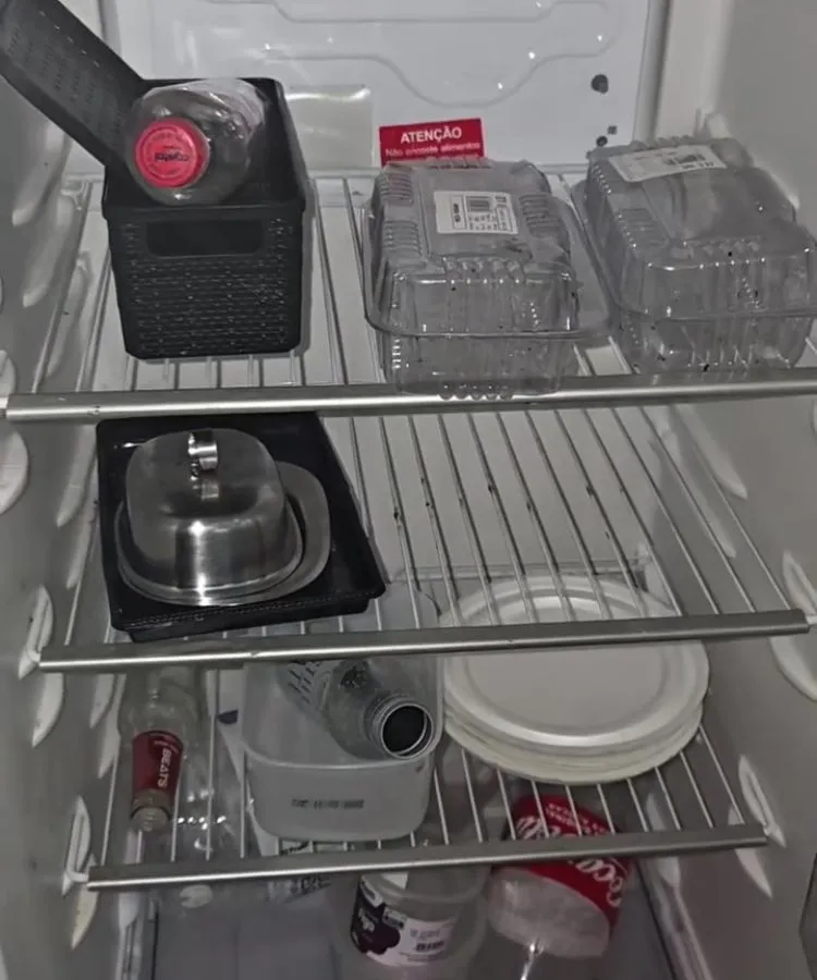 Geladeira aberta com prateleiras quase vazias, contendo recipientes plásticos, garrafa de refrigerante, potes descartáveis e uma manteigueira metálica. Alguns itens estão deitados ou fora de ordem. O eletrodoméstico branco destaca-se pela escassez de alimentos e pela organização precária.