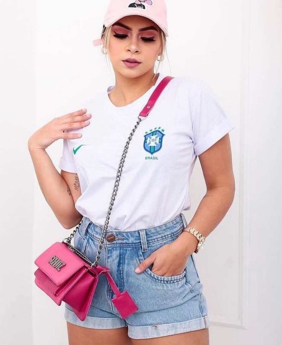 Imagem com fundo parede branca. No centro, mulher usando roupas para a copa do mundo: camiseta branca do Brasil + short jeans + bolsa pink e boné rosa.