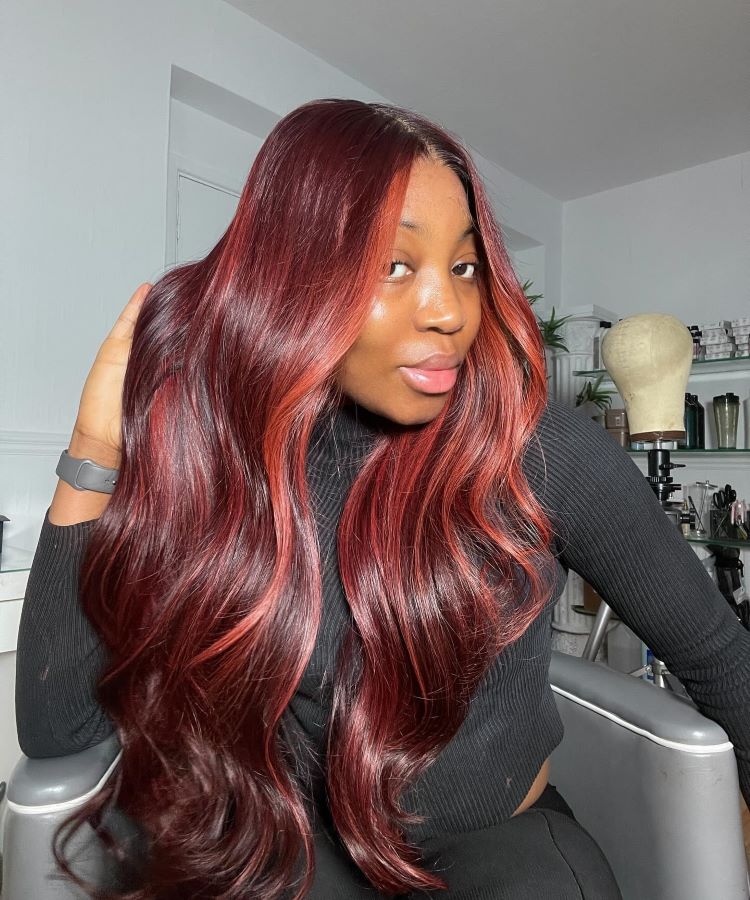 Mulher com cabelos longos e ondulados em tons vibrantes de vermelho acaju, com reflexos luminosos. Ela usa uma blusa preta de mangas compridas e está sentada em uma cadeira em um ambiente de salão, cercada por acessórios de beleza.