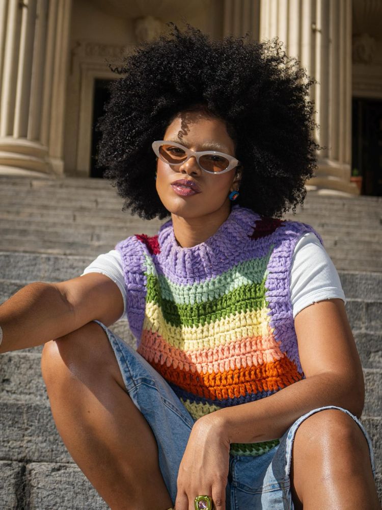Josy Ramos usando óculos gatinho arredondado com lente marrom, colete colorido, blusa branca e bermuda jeans 