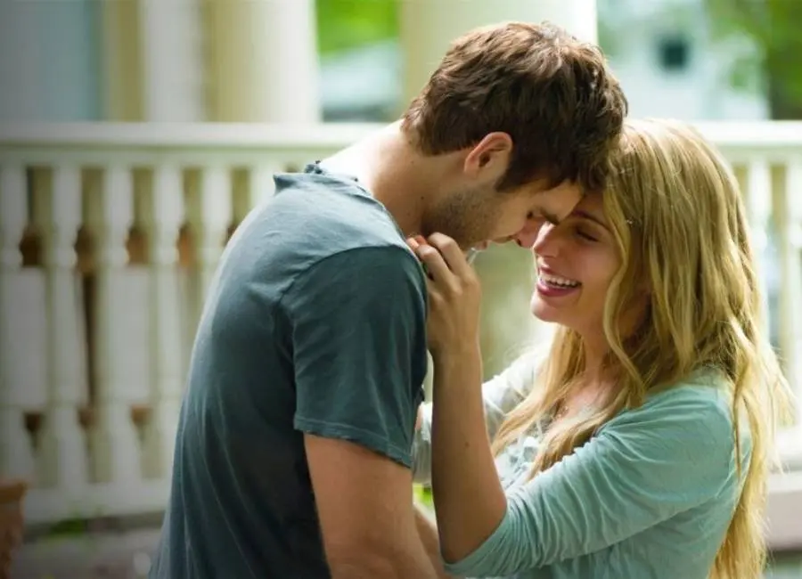 Atores do filme "A Minha Garota para Sempre" abraçados. Casal está sorridente, ela segura próximo do pescoço dele. Eles estão próximo de uma casa com sacada.