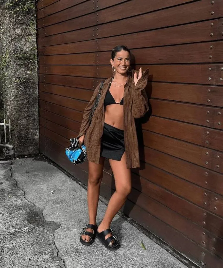 A foto mostra uma mulher com um biquini e uma saia pretos, uma camisa marrom aberta e uma sandália preta de fivelas, ela está posando apoiada em uma parede de madeira marrom escuro