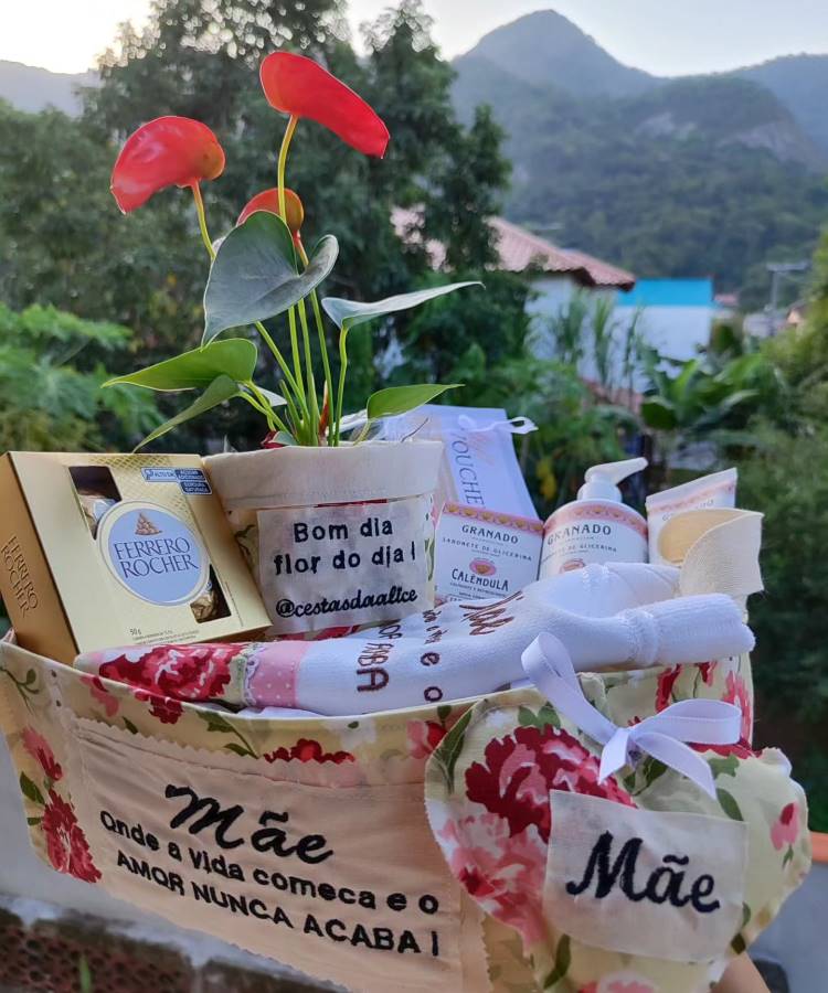 Uma cesta de presente com tema de Dia das Mães, contendo plantas, chocolates Ferrero Rocher e produtos de beleza Granado. Bordado destacando "Mãe, onde a vida começa e o amor nunca acaba." Fundo com montanhas e vegetação exuberante.





