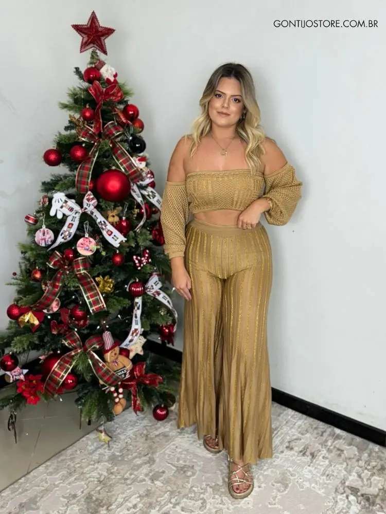 Mulher de pele clara usando cropped bege de crochê, calça dourada, ao lado de árvore de Natal