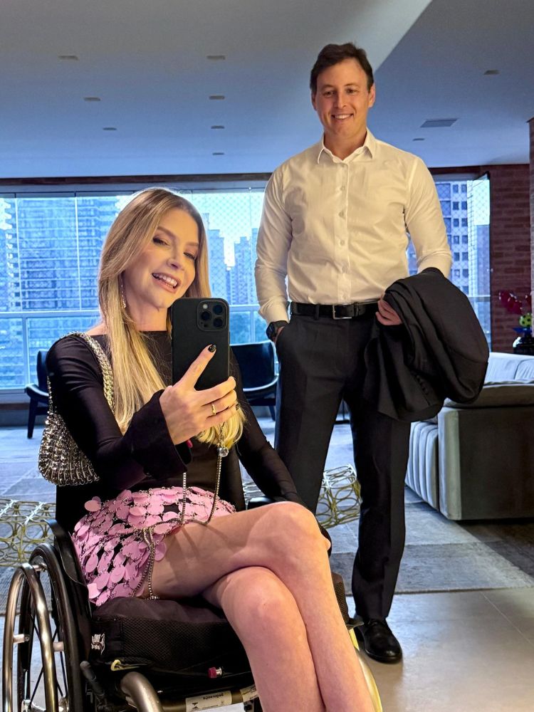Foto no espelho de Dea Schwarz de blusa preta e saia rosa e marido Jaques de camisa social branca e calça social preta