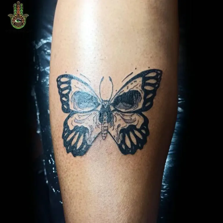 Tatuagem de borboleta com estampa de caveira feita em traços grossos na canela de um homem de pele clara. 