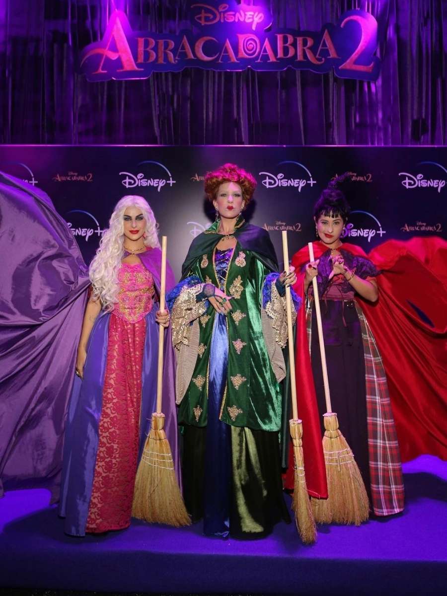 Imagem em fundo do painel da festa Halloween Sephora. Na parte superior lê-se em abóbora: "Disney Abracadabra 2". Mais abaixo, contem as logos do filme da Disney espalhadas em um painel preto. No centro da foto e mais abaixo, Nah Cardoso, Lore Improta e Thaynara OG fantasiadas das Irmãs Sanderson do filme. Nah veste vestido roxo e rosa com capa e peruca longa branca, Lore veste vestido verde e azul com capa e peruca ruiva, Thaynara verde vestido roxo e vermelho com peruca preta. As três seguram vassouras de bruxa. 