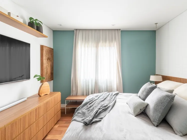Quarto do casal com cama king size. Em frente, móvel com gavetas, TV na parede e prateleira em cima com vasos, folha de costela-de-adão e mais caixas. Na parede, um quadro decora.