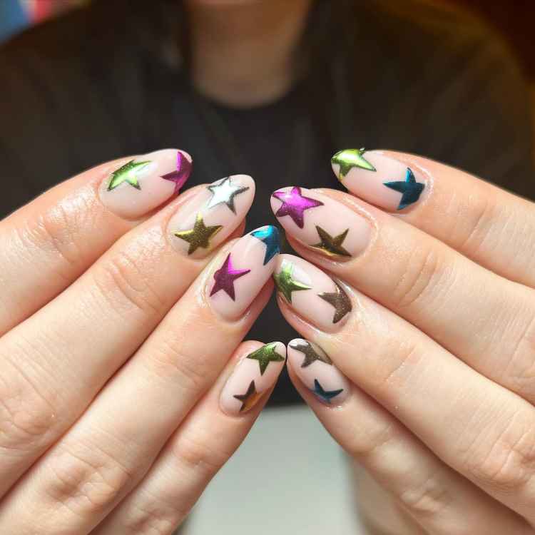 Unhas longas com esmalte nude decoradas com estrelas metálicas em cores vibrantes como roxo, verde, azul e dourado. As mãos estão entrelaçadas, destacando o design alegre e moderno das unhas contra o fundo neutro, criando um efeito visual impactante.
