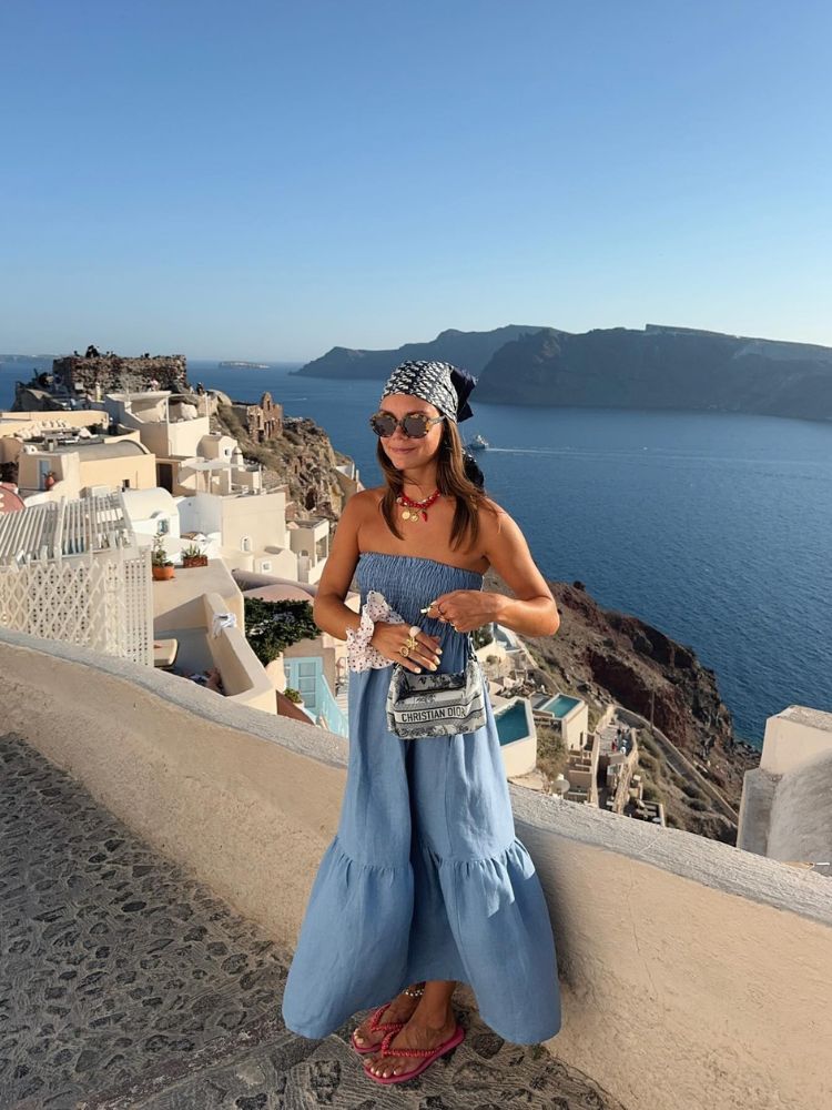 Mulher de pele clara usando vestido azul e bandana branca e preta com mar ao fundo 