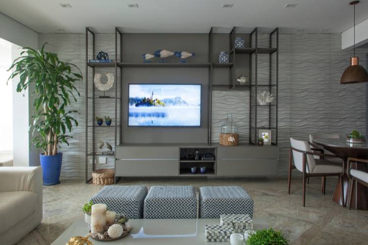 Sala de estar em tons de cinza com estante vazada em algumas partes e fechada em outras. Ela comporta a televisão e diversos itens decorativos. 