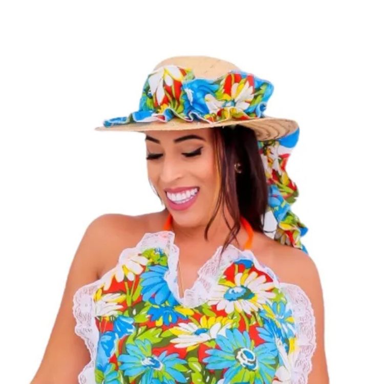 Uma mulher sorridente usa um chapéu de festa junina de palha com uma faixa floral azul, vermelha e branca e babados. Ela também veste uma blusa com estampa floral colorida e detalhes de renda branca.