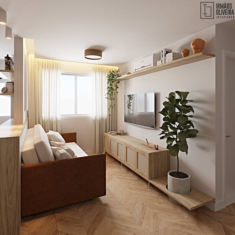 A imagem mostra o interior de uma sala de estar moderna e bem iluminada. Há um sofá marrom claro com almofadas brancas no canto esquerdo da imagem. Uma televisão plana está montada na parede, cercada por prateleiras e armários de madeira clara. Uma planta verde em um vaso branco está posicionada à direita da televisão. Cortinas amarelas claras cobrem uma janela ao fundo, permitindo que a luz natural entre na sala. Diversos objetos decorativos estão dispostos nas prateleiras acima da televisão. A sala é espaçosa, com paredes brancas e um piso de madeira clara. A imagem tem uma inscrição “IRMÃOS OLIVEIRA INTERIORES”.
