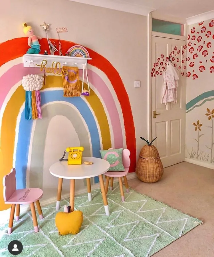 Quarto infantil alegre com parede pintada de arco-íris, mesa branca com cadeiras rosa e verde, telefone retrô amarelo, prateleira decorada com brinquedos, almofadas coloridas, cesto em formato de abacaxi, tapete verde geométrico e detalhes lúdicos.
