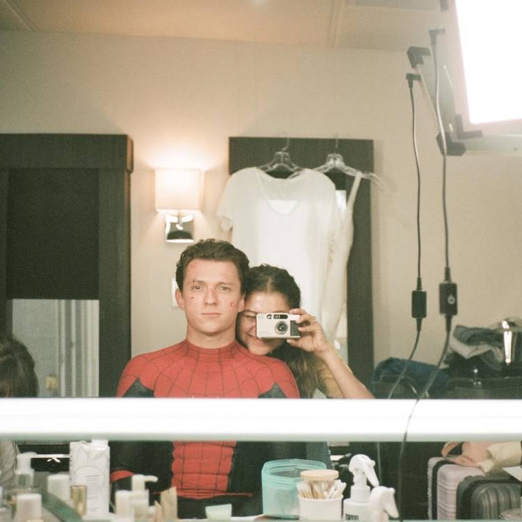 Tom Holland e Zendaya tiram uma selfie no espelho. Tom, usando o traje do Homem-Aranha, parece sério. Zendaya, por trás, esconde o rosto atrás de uma câmera, capturando o momento. A cena é dos bastidores. Famosas que estão noivas