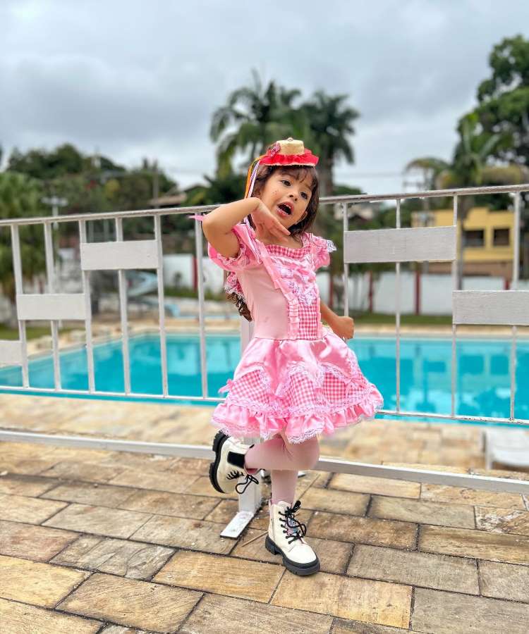 Menina usando vestido de quadrilha rosa com coturno branco e, como acessório, um mini chapéu de palha no cabelo.
