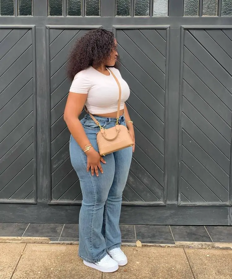 Uma mulher de cabelo encaracolado e castanho está em frente a uma porta preta. Ela veste um top branco curto, calça jeans flare de cintura alta e tênis branco. Ela está usando joias douradas e uma bolsa bege.