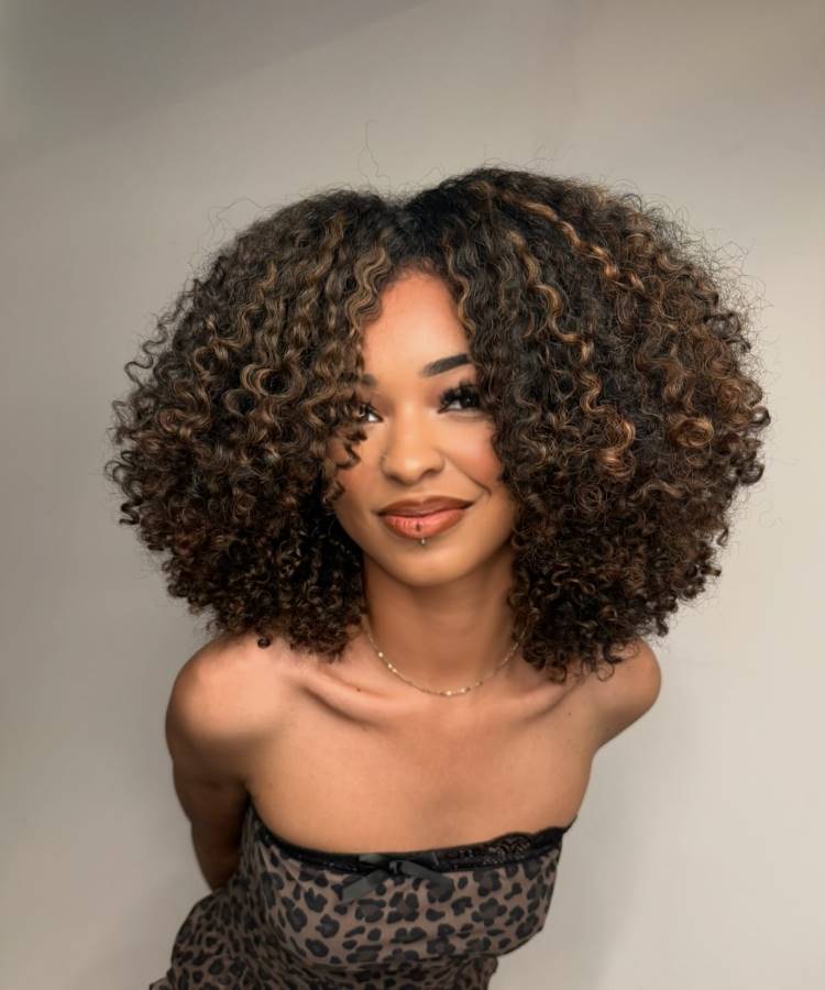 A imagem mostra uma jovem com cabelo castanho iluminado, volumoso e cacheado, sorrindo levemente. Ela usa um top tomara que caia com estampa de oncinha e tem um piercing no lábio. O fundo é neutro e destaca a modelo e seu cabelo.







