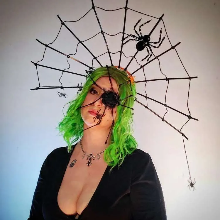 Mulher de pele clara e cabelo verde usando tiara de teia de aranha, maquiagem de olho machucado e vestido preto 