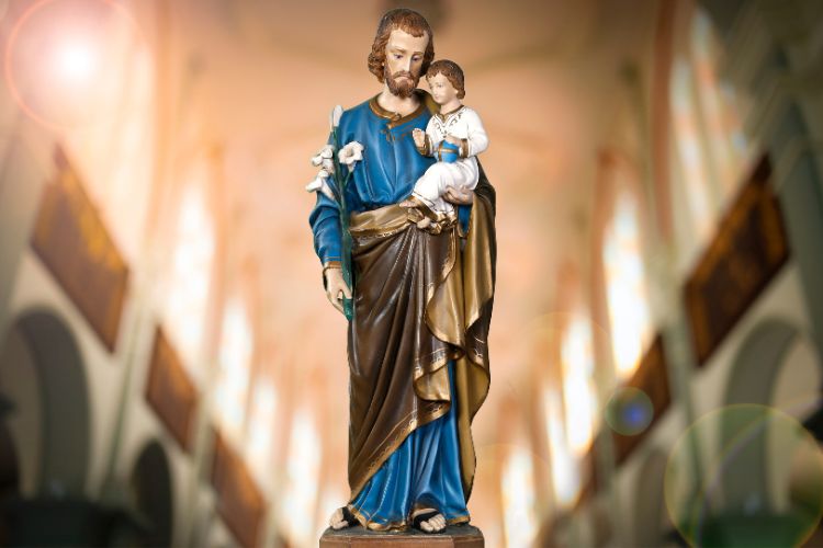 Imagem de uma estátua de São José segurando o Menino Jesus nos braços. Ele veste túnica azul com manto dourado, segurando lírios, símbolo de pureza. O fundo desfocado mostra o interior de uma igreja iluminada.