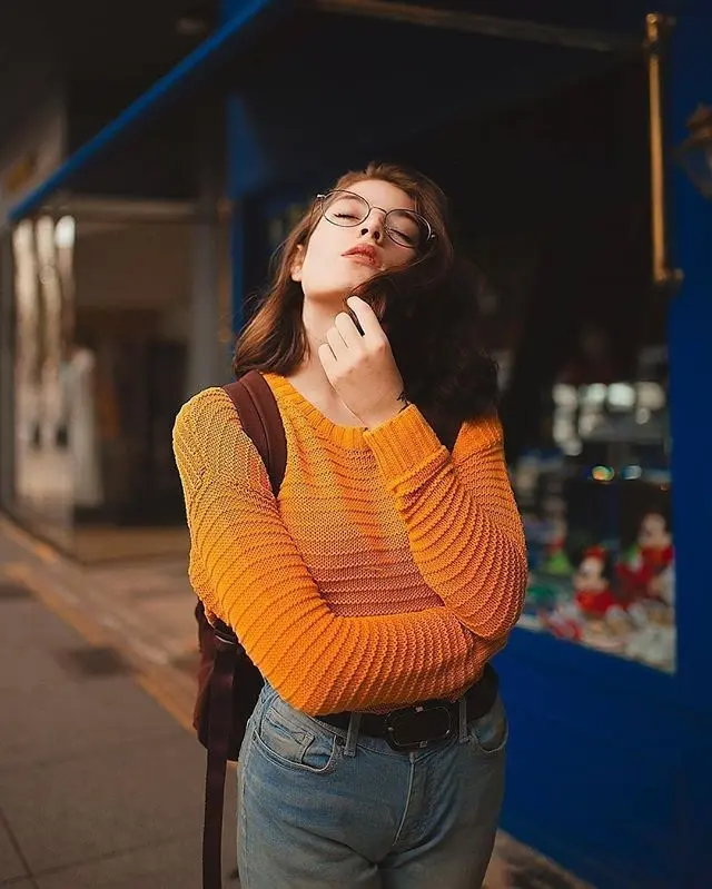 Frase para foto pensativa usa óculos de grau e veste calça jeans com cinto preto e camisa de tricô laranja