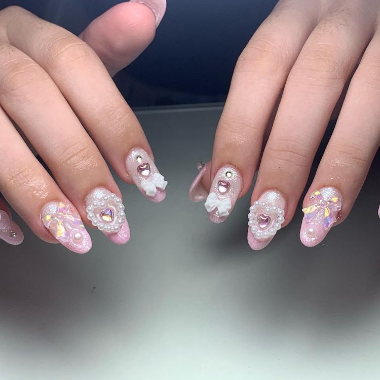 Pessoa de pele clara com unha estilo coquette, no formato almond, com pedrarias e laço, tudo em rosa claro 