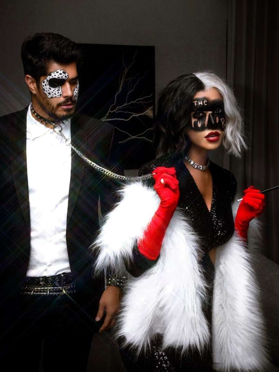 Imagem em fundo de parede com quadro e cortina e Rica de Marré e Abrão Meron na frente. Casal está fantasiado, ela de Cruella e ele de Dálmata. Ela veste vestido preto, luvas vermelhas, com manto de pelos brancos nos braços, além de peruca preta e branca e maquiagem preta com "The Future" escrito no rosto. Ele veste terno e possui maquiagem imitando as pintinhas de um dálmata no rosto.