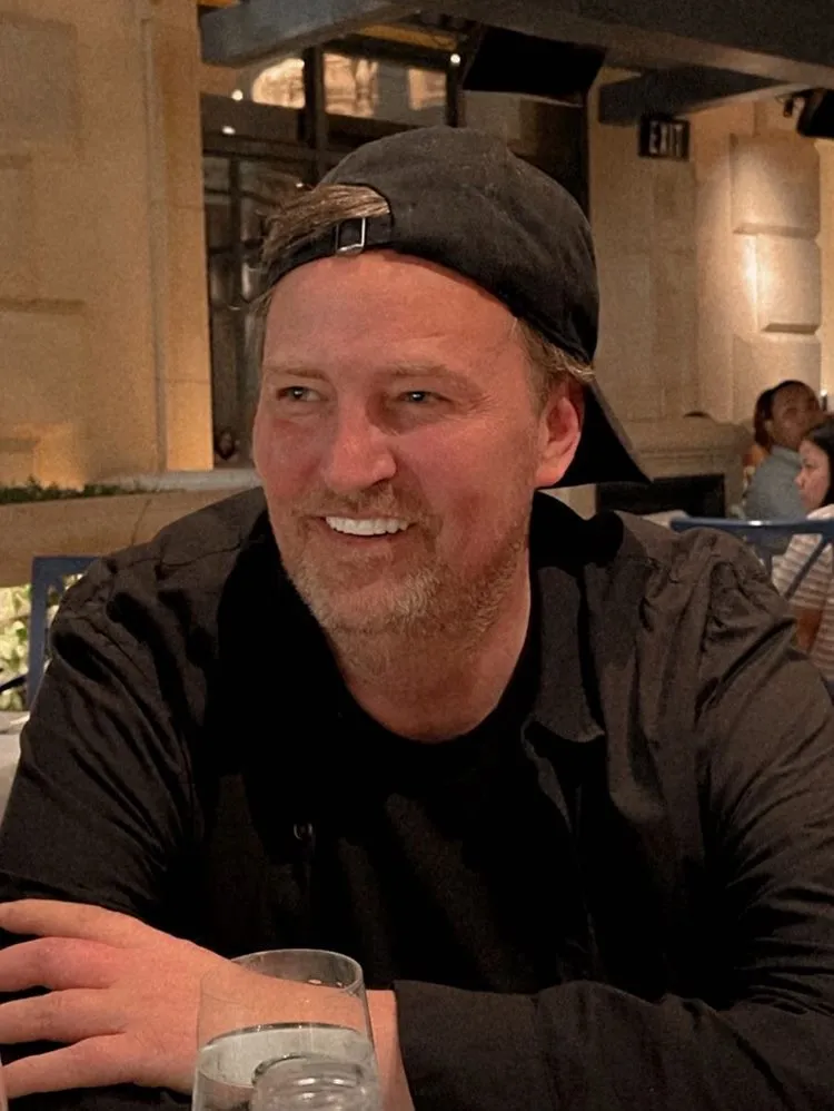 Ator Matthew Perry e foto sorridente, sentado em restaurante, usando boné preto, camisa e camiseta pretos. Em sua frente uma taça com água. Notícia retrospectiva 2023