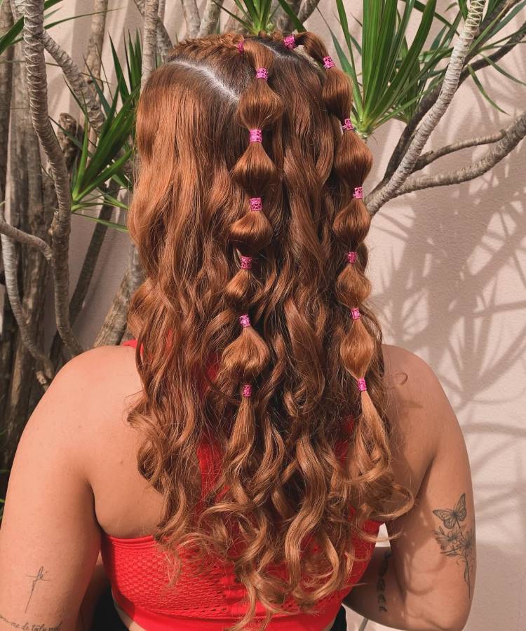 A imagem mostra uma mulher de costas com cabelos ruivos cacheados, presos em um penteado com bolhas, decorado com elásticos rosa. Ela veste um top vermelho e exibe uma tatuagem de borboleta no braço esquerdo.