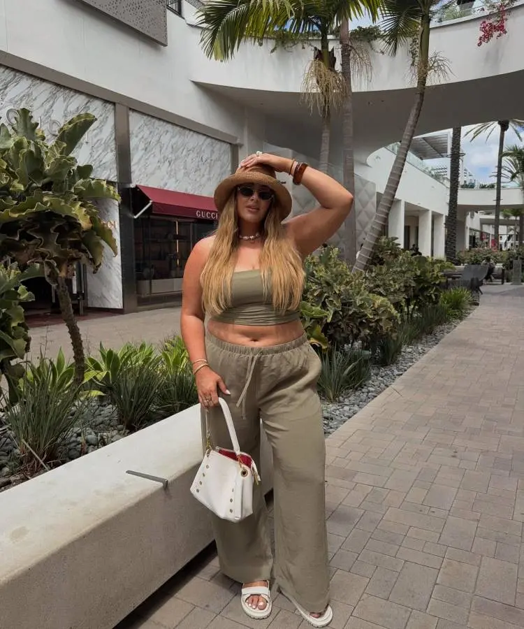 Uma mulher loira posa casualmente em um shopping ao ar livre, segurando seu chapéu de palha. Ela usa um conjunto verde-oliva de calça larga e top, chinelos brancos e uma bolsa branca com detalhes dourados.
